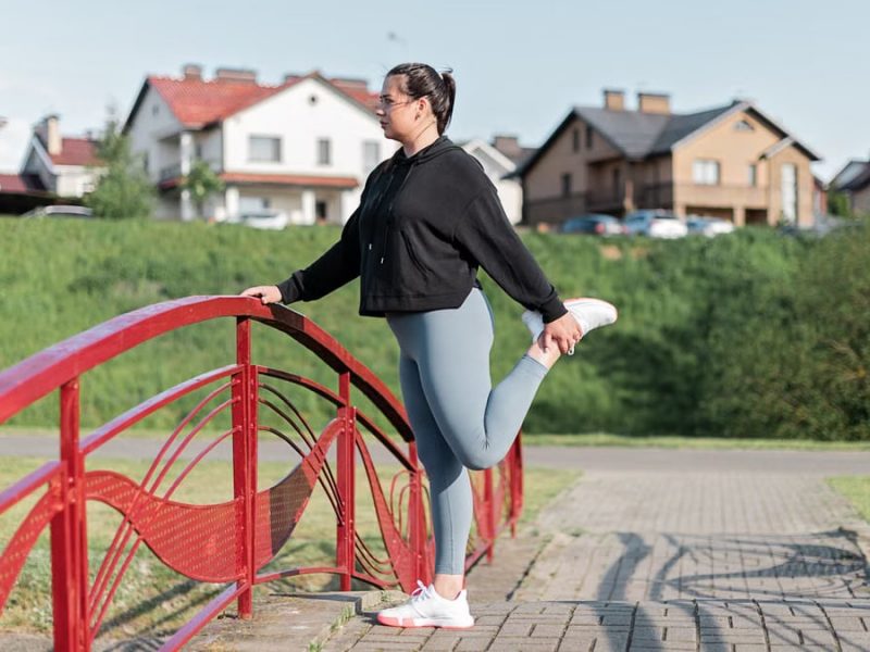 A woman in active wear performs a stretching exercise on a red bridge in a suburban park setting.