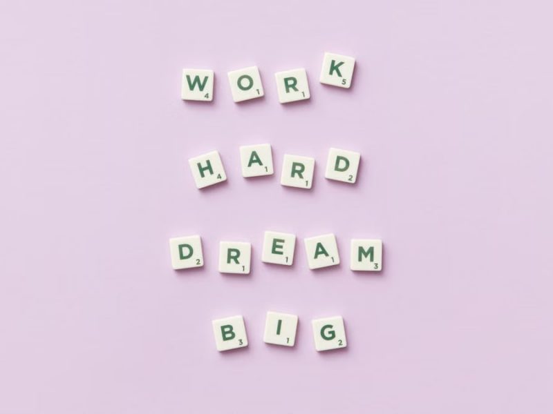 Scrabble tiles spell 'Work Hard Dream Big' on a pastel purple surface, top view.